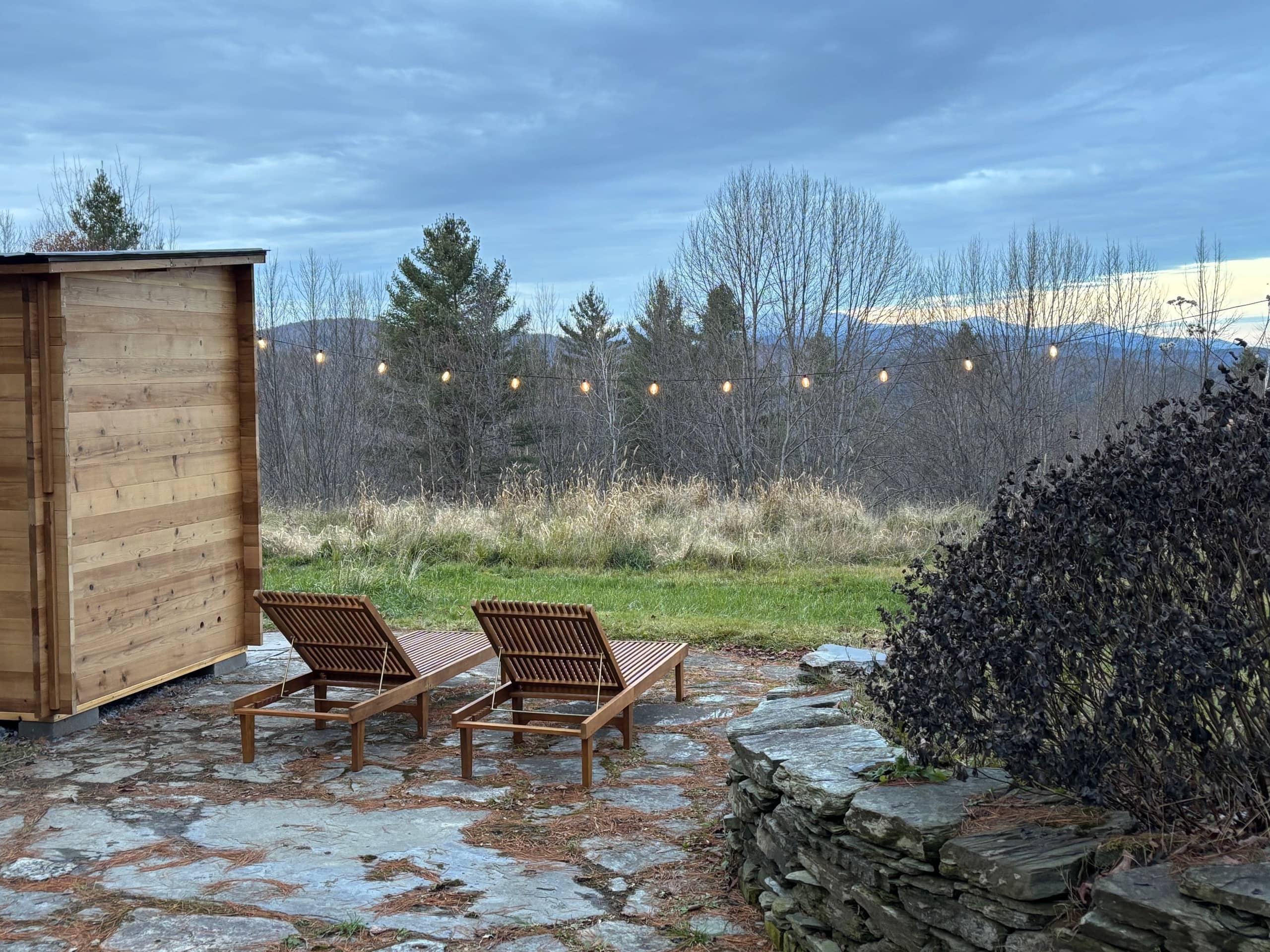 Cedar outdoor sauna beside a stone patio with teak loungers, string lights, and mountain views